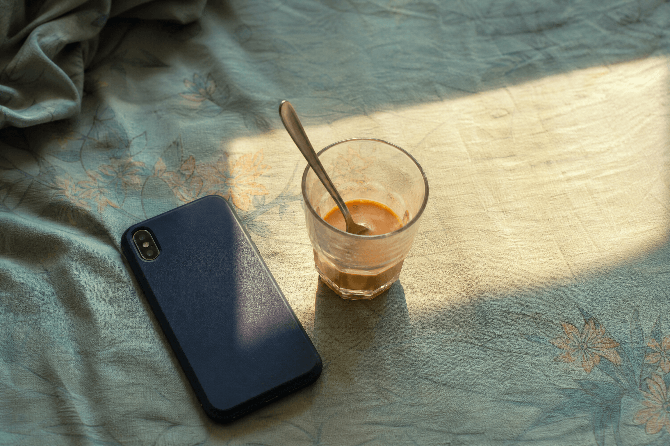 A phone lying face-down on a cotton bedsheet beside a half-drunk glass of chai, afternoon light across the fabric